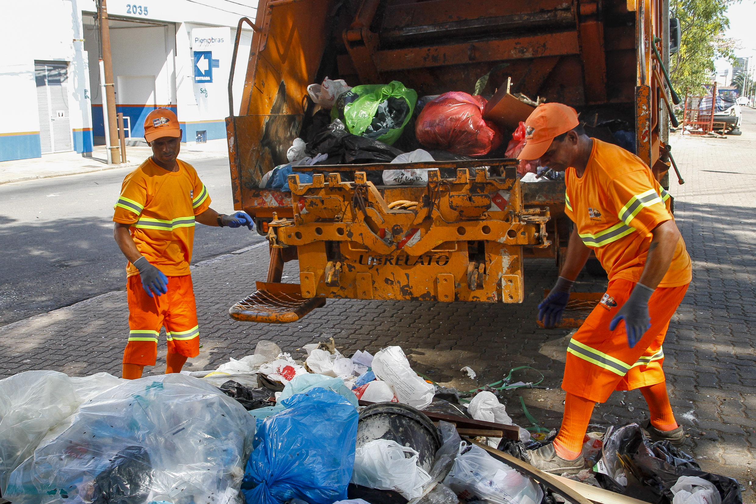 Dois garis, vestidos com unifome laranja, fazem o serviço de coleta de lixo junto a um caminhão próprio para o recolhimento.