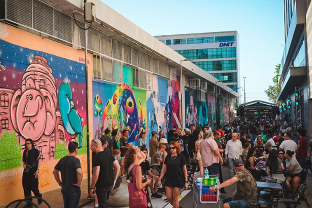 Rua movimentada com muitas pessoas, murais coloridos nas paredes e clima de confraternização em um dia ensolarado.